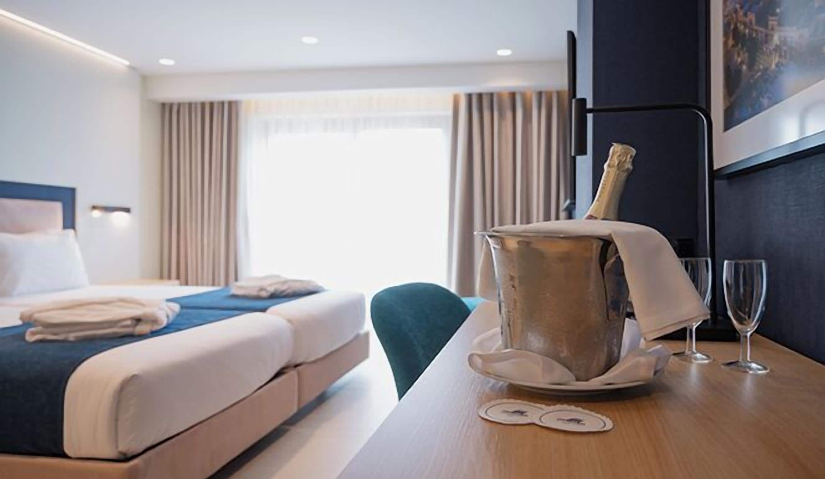 Ein Hotelzimmer mit einem Bett, einer Minibar, zwei Weingläsern und einem Tisch mit zwei Untersetzern.
