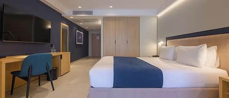 A modern hotel room with a large bed, a desk, a chair, a TV, and a wardrobe.