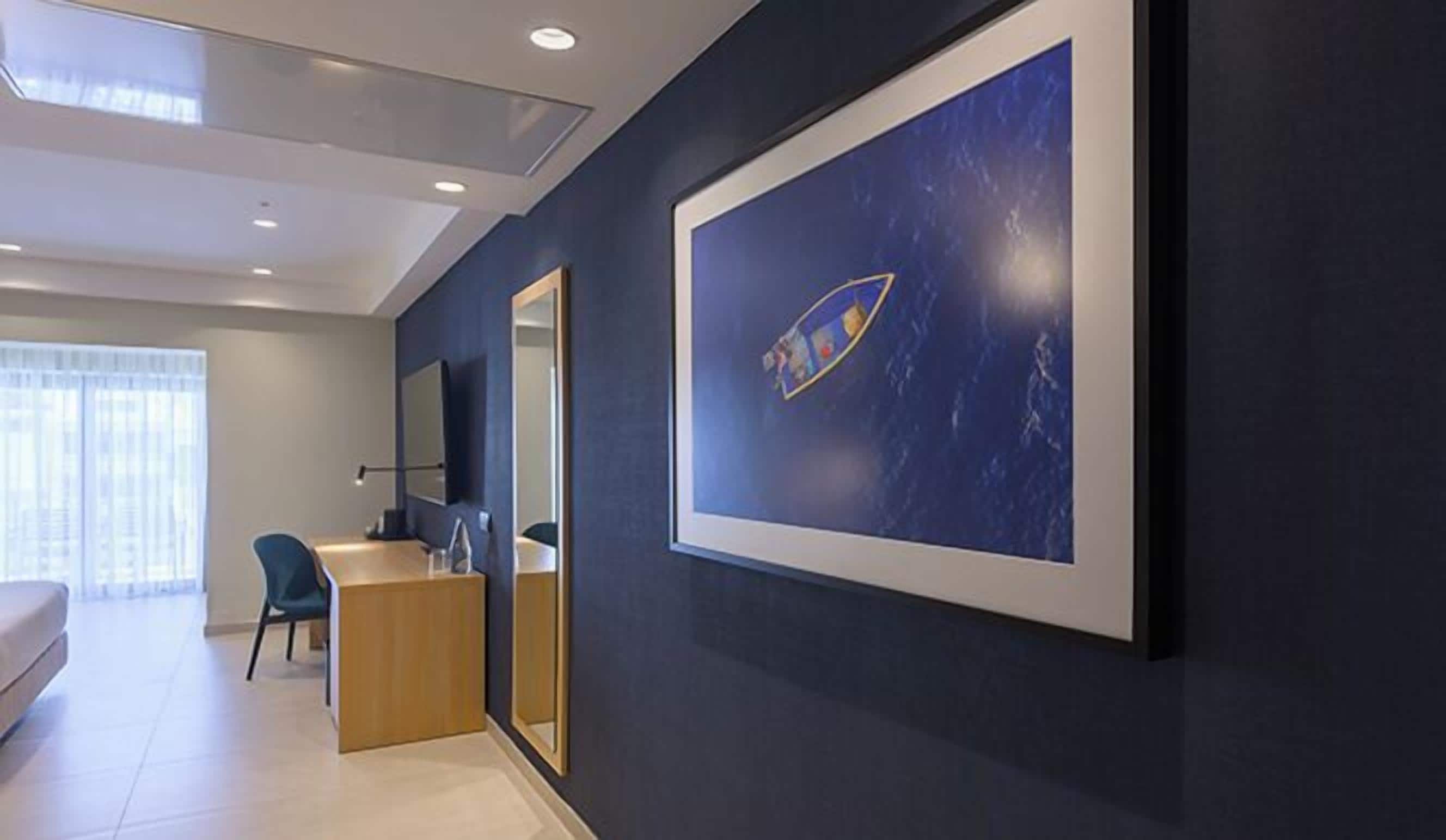 Habitación de hotel moderna con una gran obra de arte enmarcadas de un barco sobre fondo azul, un escritorio de madera, una silla y un espejo grande.