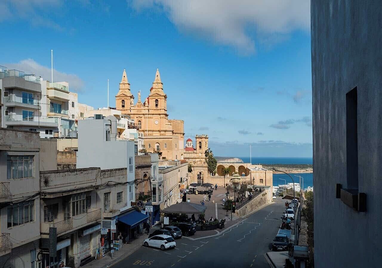 A coastal town with a prominent church featuring two towers, a street lined with parked cars, and a view of the sea.