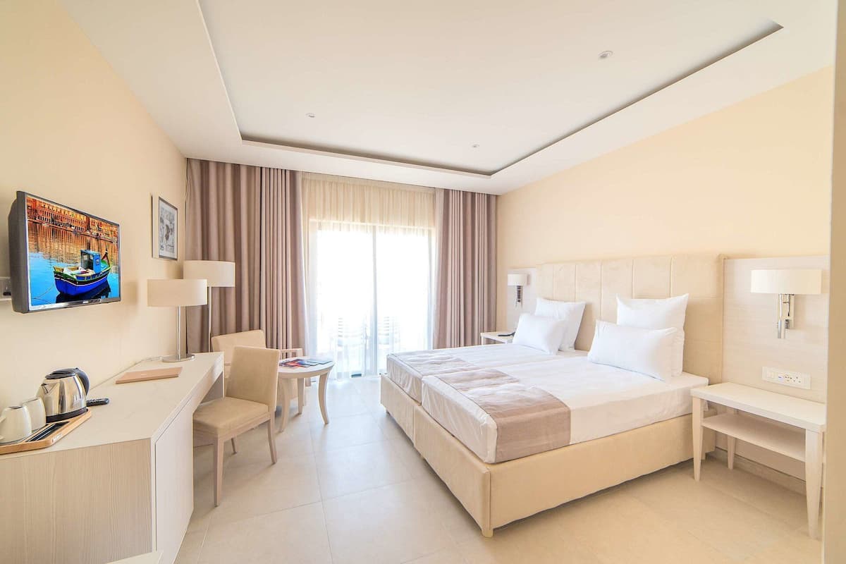 A hotel room with a bed, bedside tables, a desk with a chair, a TV, and a window with curtains.