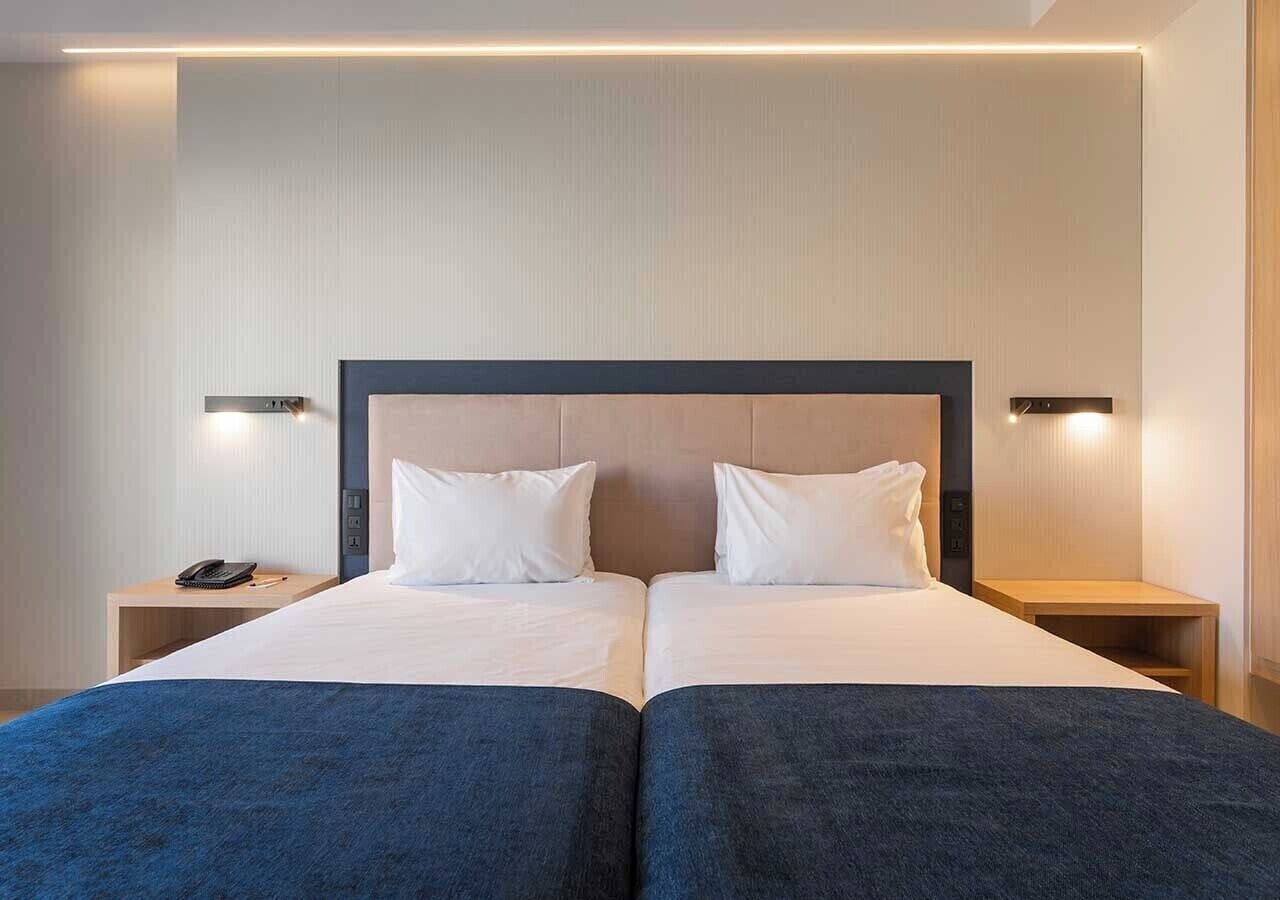 A double bed with a dark blue bedspread, two white pillows, and a wooden headboard. There are two bedside tables, one on each side of the bed, with a black telephone on the left table.