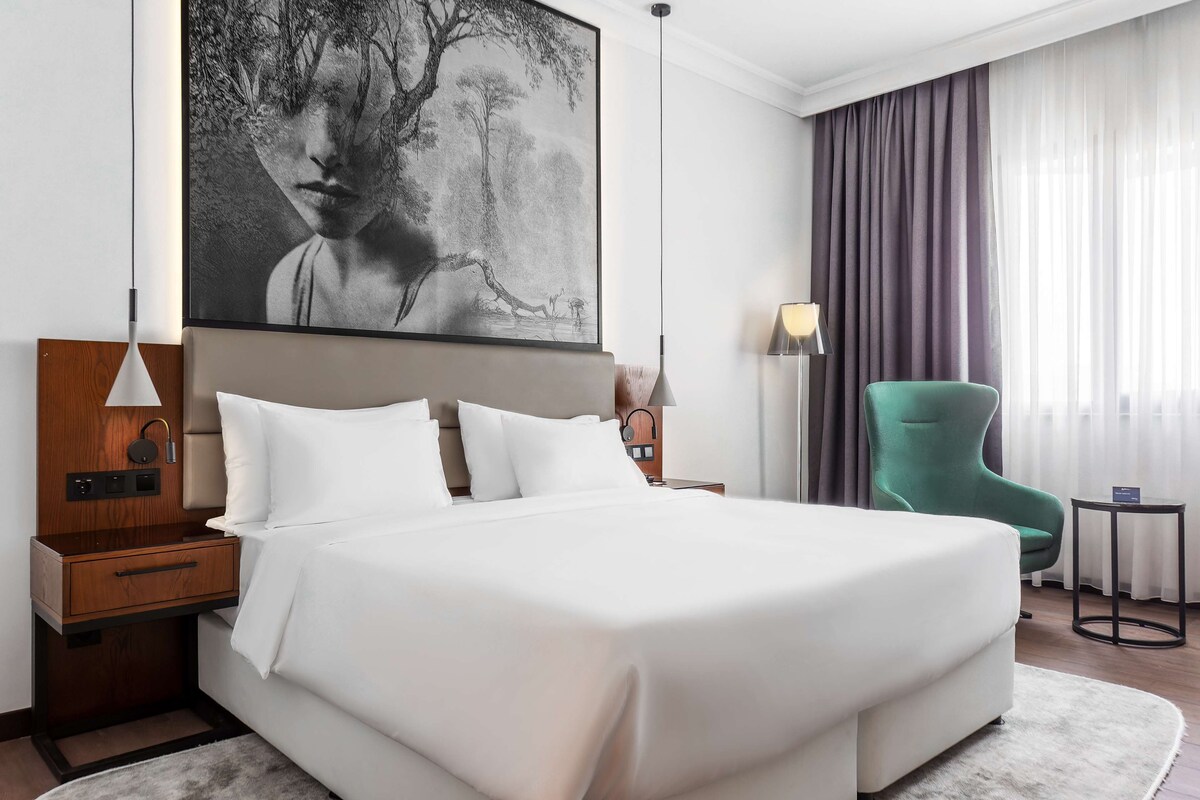 Una habitación de hotel moderna con una cama grande, una mesita de noche con lámpara, un sillón verde y un cuadro abstracto de gran tamaño en la pared.