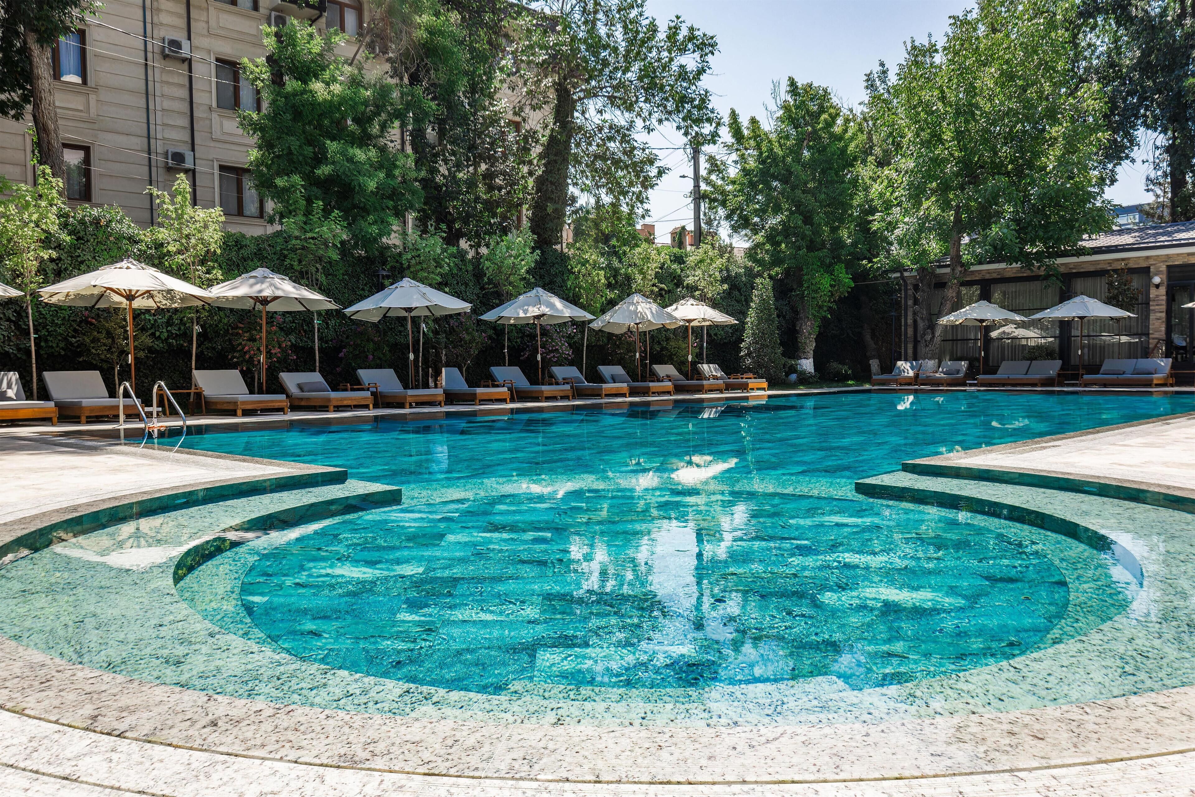 A swimming pool with lounge chairs and umbrellas, surrounded by trees and a building.