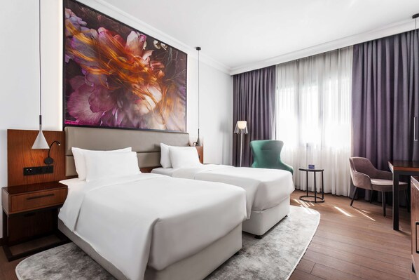 Quarto de hotel com duas camas, uma grande obra de arte floral na parede, uma mesa de trabalho com cadeira e vista para o exterior pela janela.