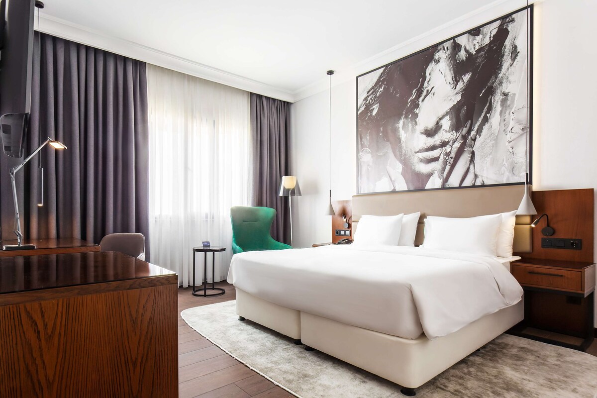 Una habitación de hotel con una cama grande, un escritorio, una silla verde y un cuadro abstracto de gran tamaño en la pared.