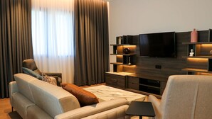 Una sala de estar moderna con sofá, sillón y mesa de centro. Hay un televisor de pantalla plana montado en la pared, y una mesita con objetos decorativos.