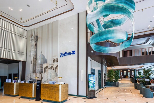 A modern hotel lobby with a reception desk, a large decorative light fixture, and a mural of a historical building.