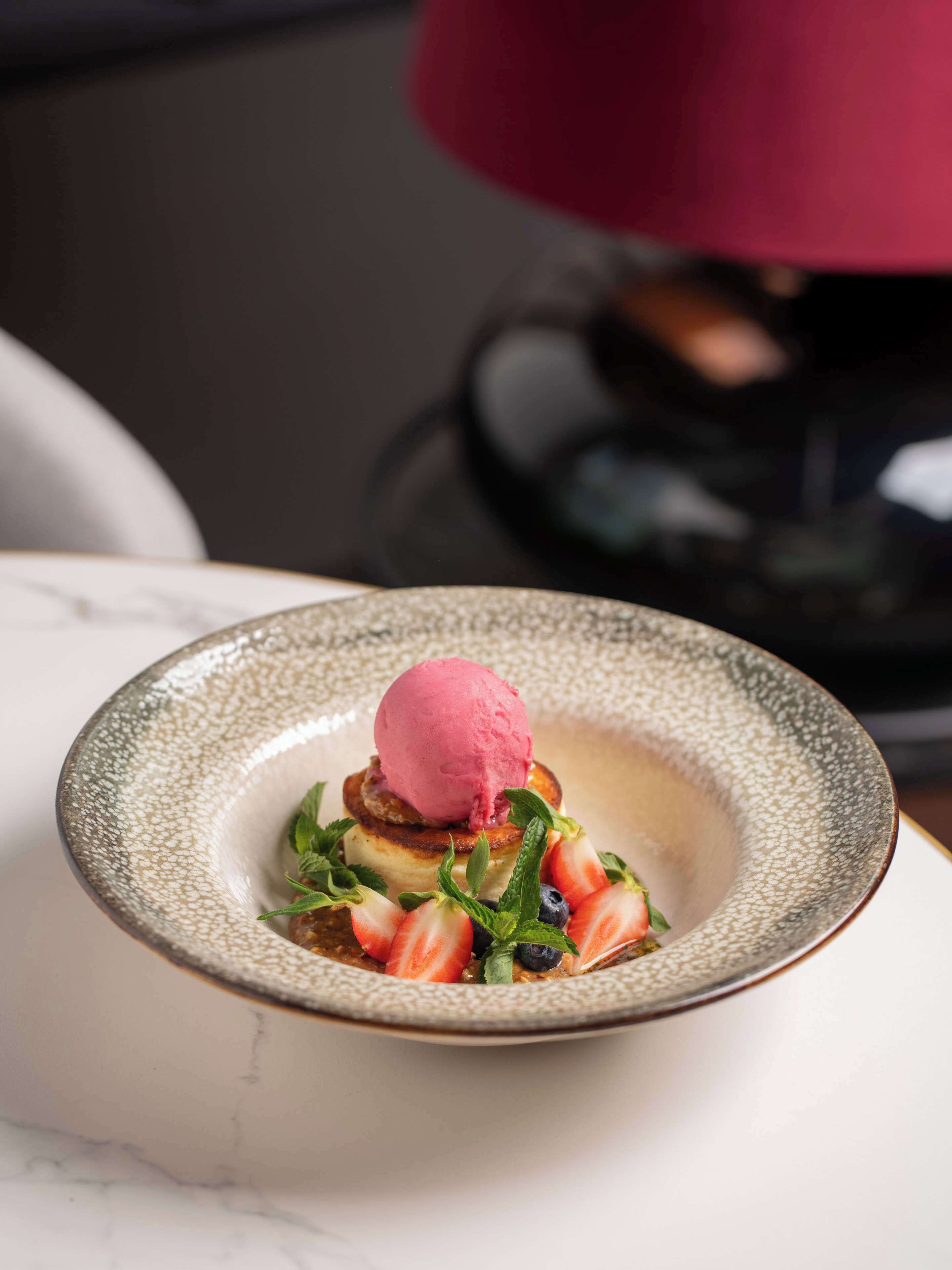 A dessert with a scoop of pink ice cream on top of a stack of pancakes, garnished with fresh berries and mint leaves, served in a textured bowl.