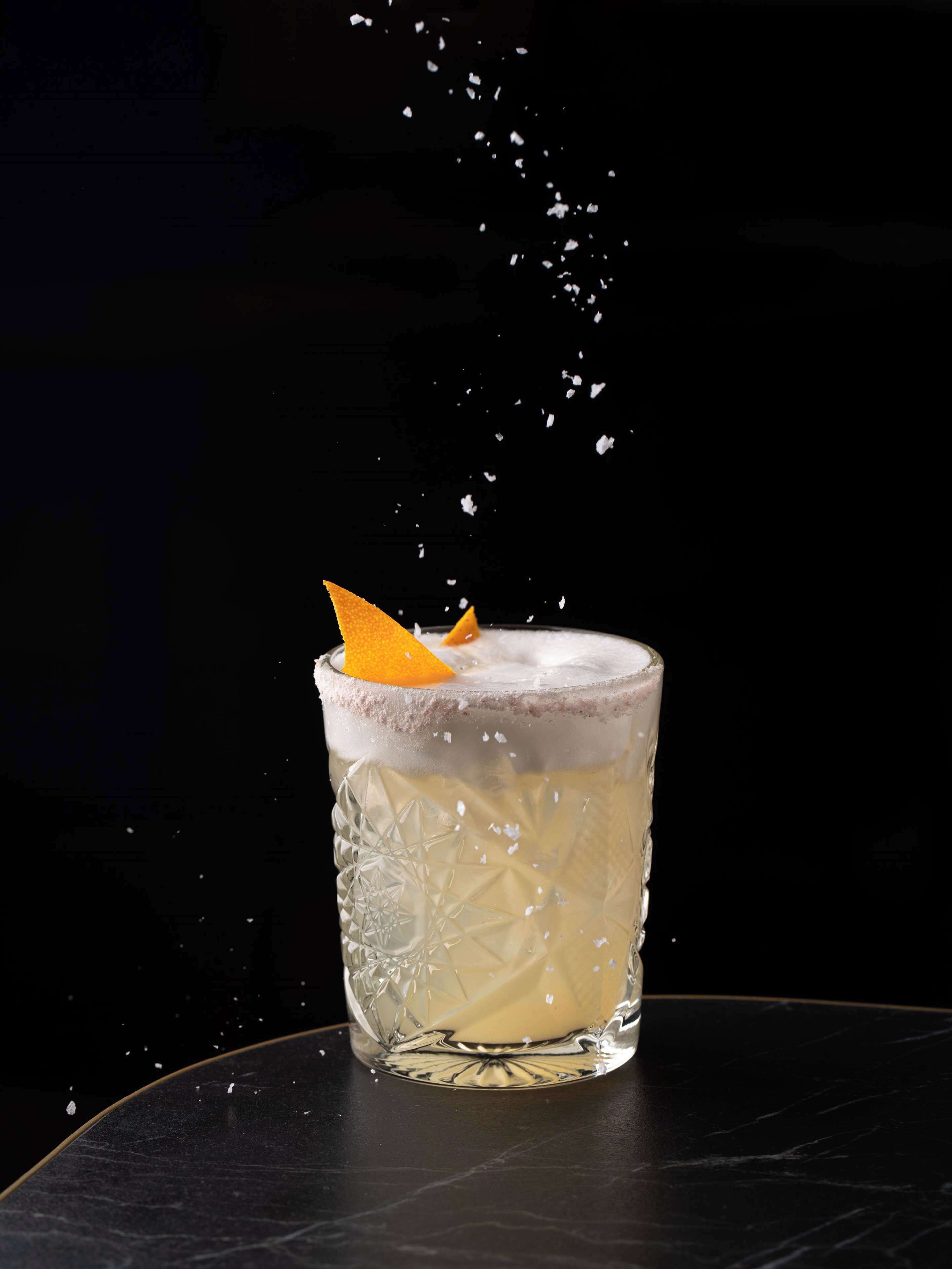Un verre transparent orné d’un motif texturé, contenant une boisson surmontée d’une mousse et décorée d’une tranche d’orange, le tout présenté sur un fond sombre.