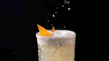 Un vaso transparente con diseño texturizado, conteniendo una bebida con espuma y una decoración de naranja, sobre un fondo oscuro.
