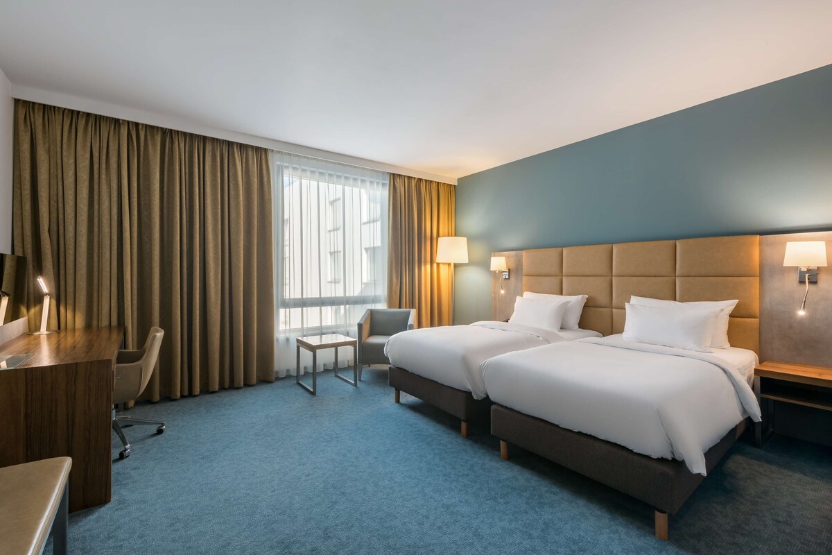 Habitación de hotel con dos camas, un escritorio, una silla y un ventanal grande con cortinas.