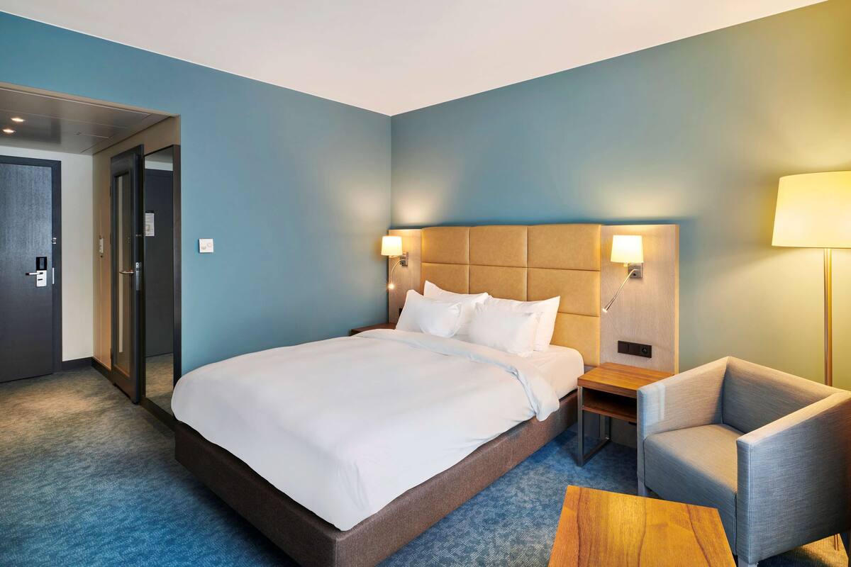 A hotel room with a bed, a chair, a desk, and a lamp.