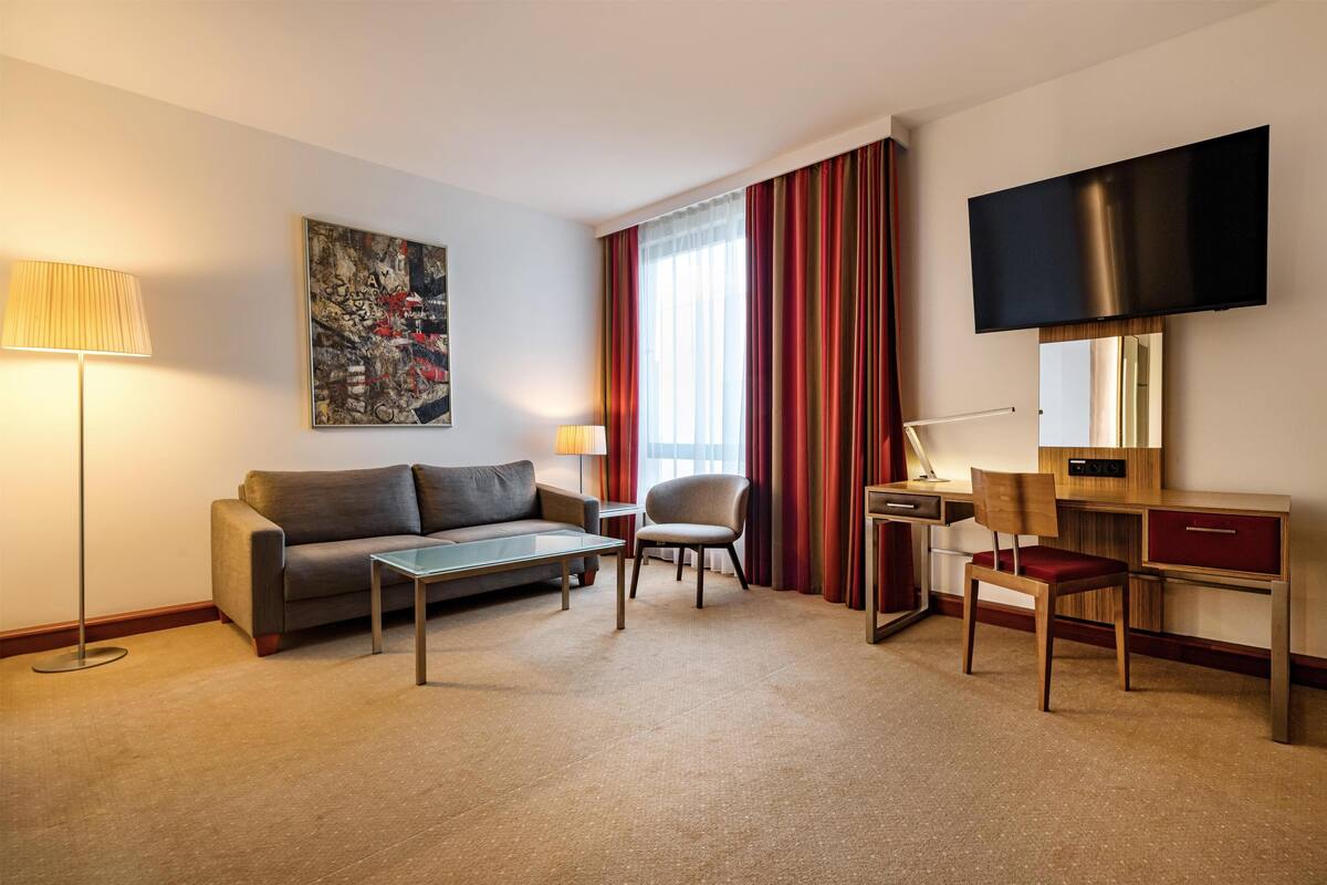 A hotel room with a sofa, a chair, a desk, a flat-screen TV, a lamp, and a painting on the wall.