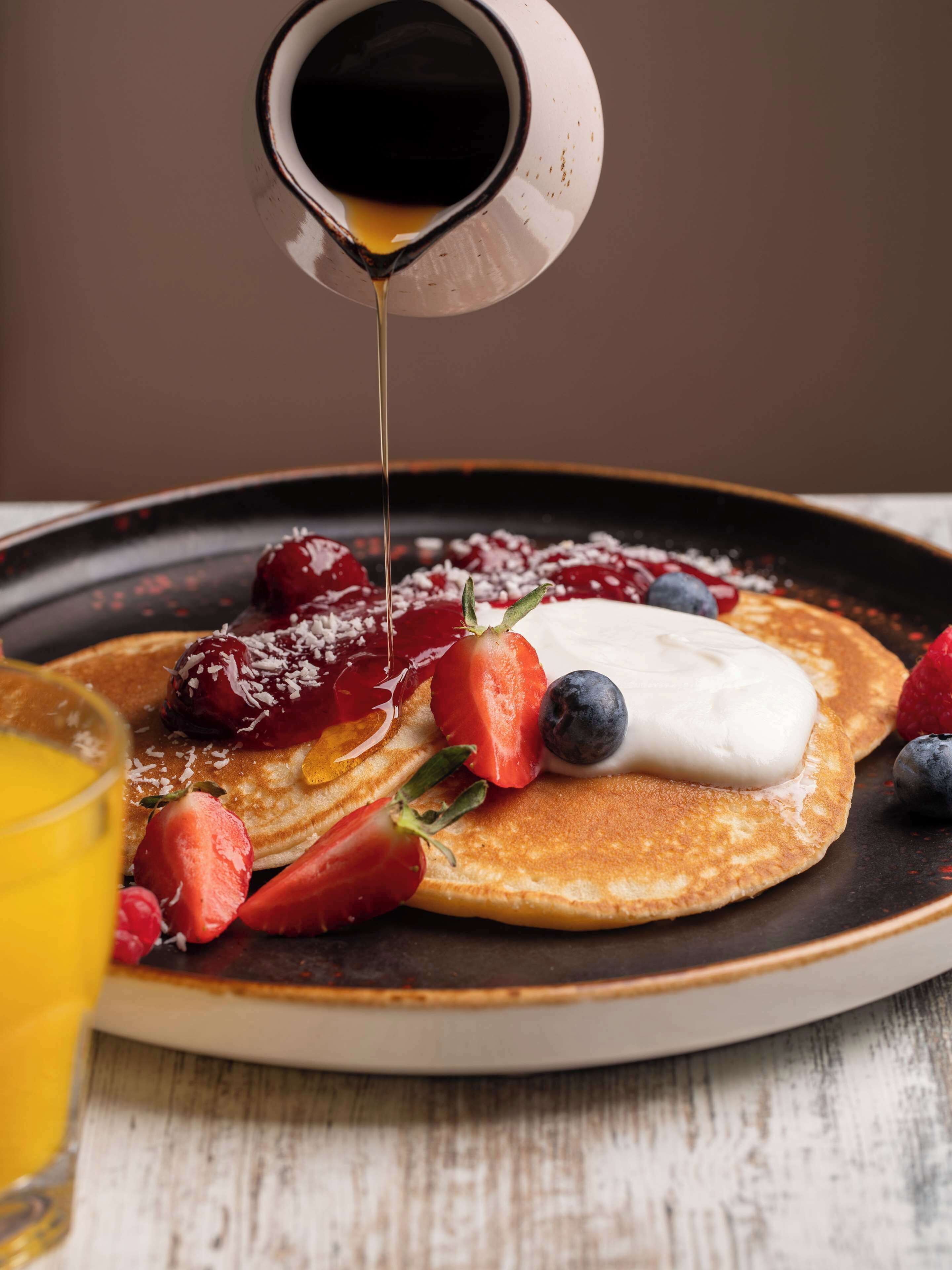 A plate of pancakes with strawberries, blueberries, and a dollop of cream, with syrup being poured over it.