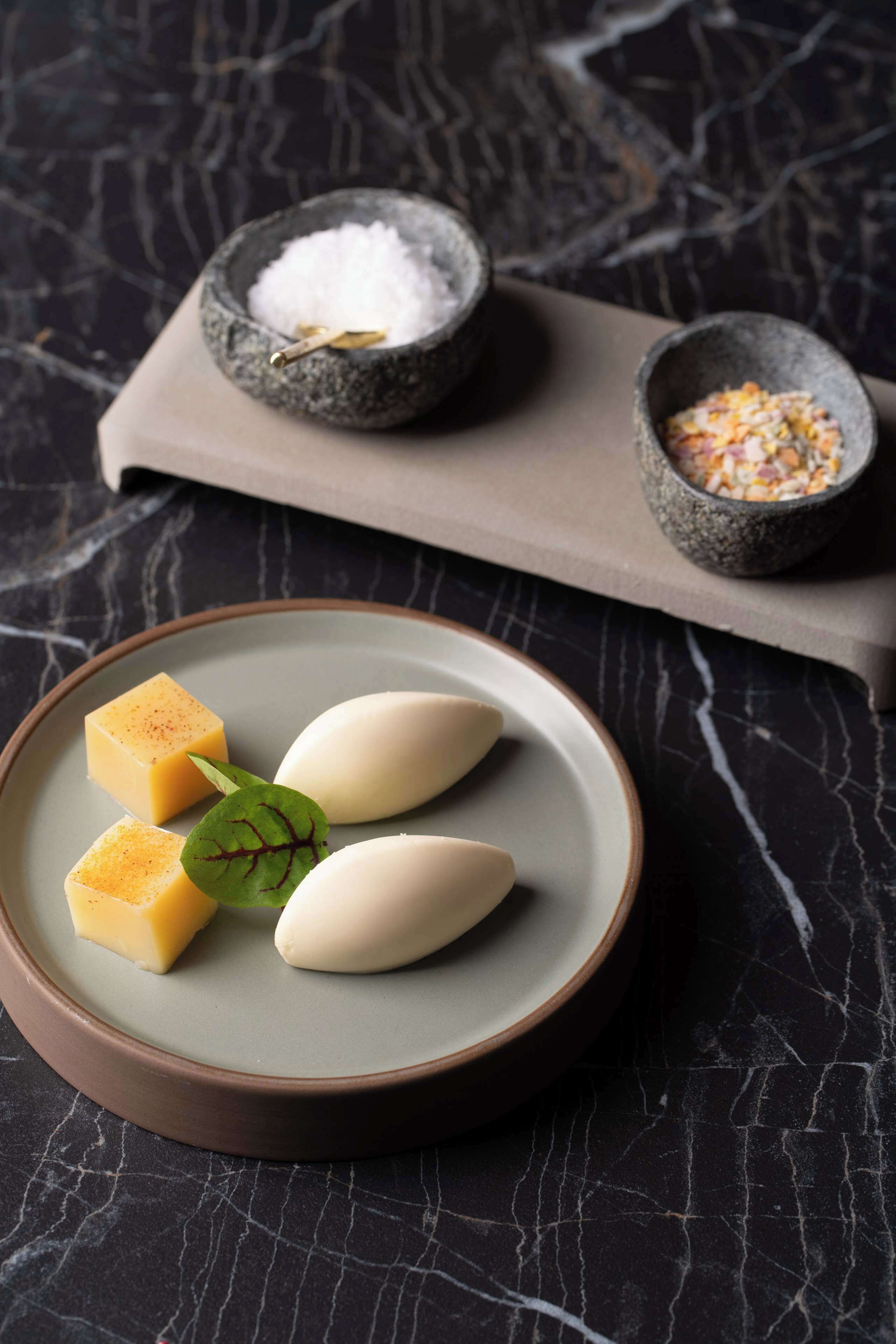 Un plato con tres cubos de queso y dos elementos ovalados blancos, posiblemente queso, sobre una superficie de mármol oscuro.