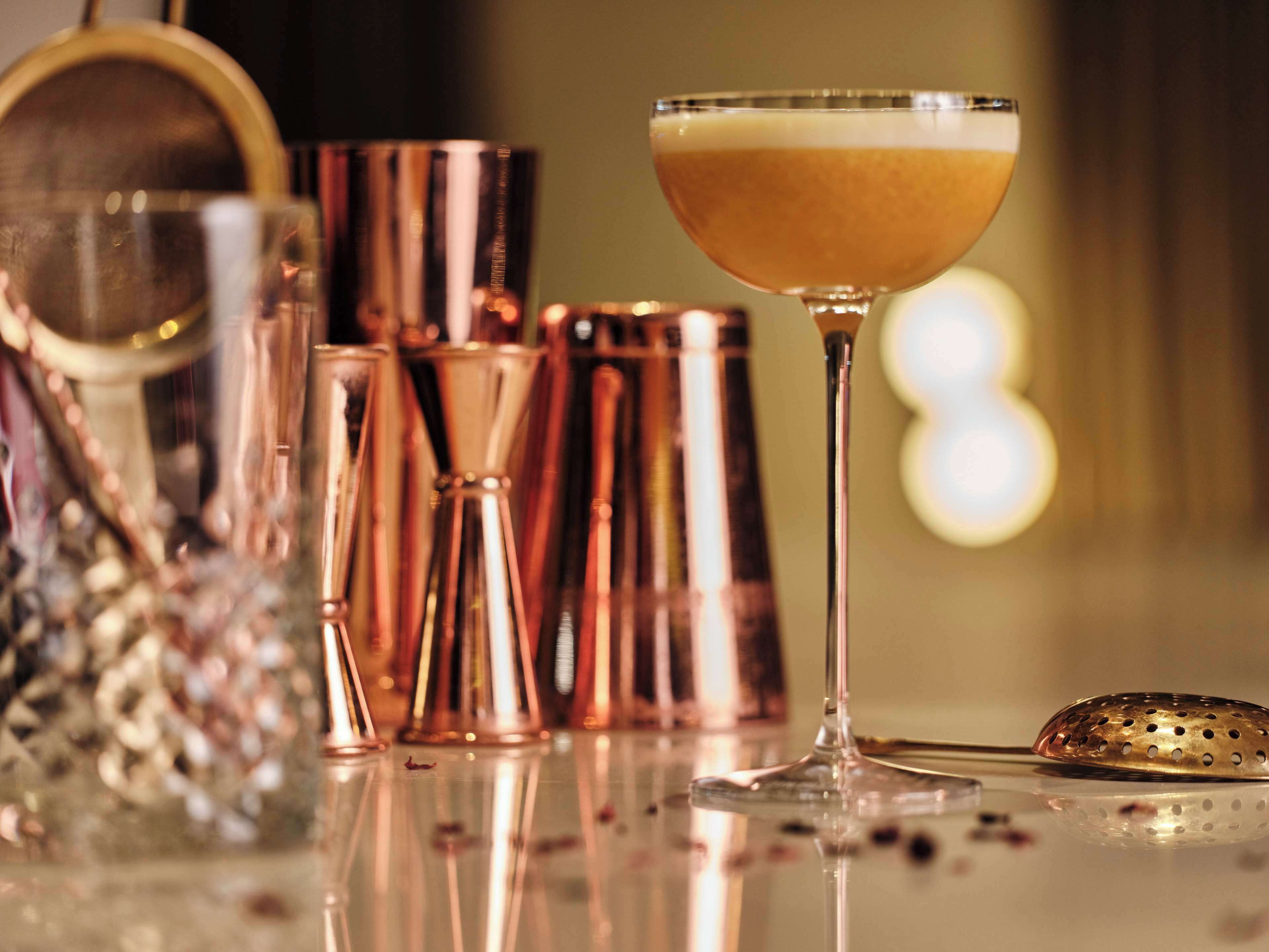 A cocktail glass with a layered drink, surrounded by copper-colored cocktail shakers and a strainer on a reflective surface.