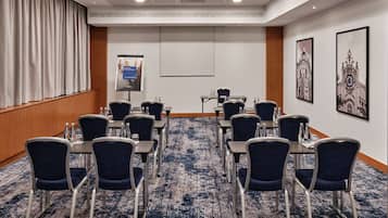 A conference room with a carpeted floor, rows of chairs, tables, a projector screen, and two framed pictures on the wall.