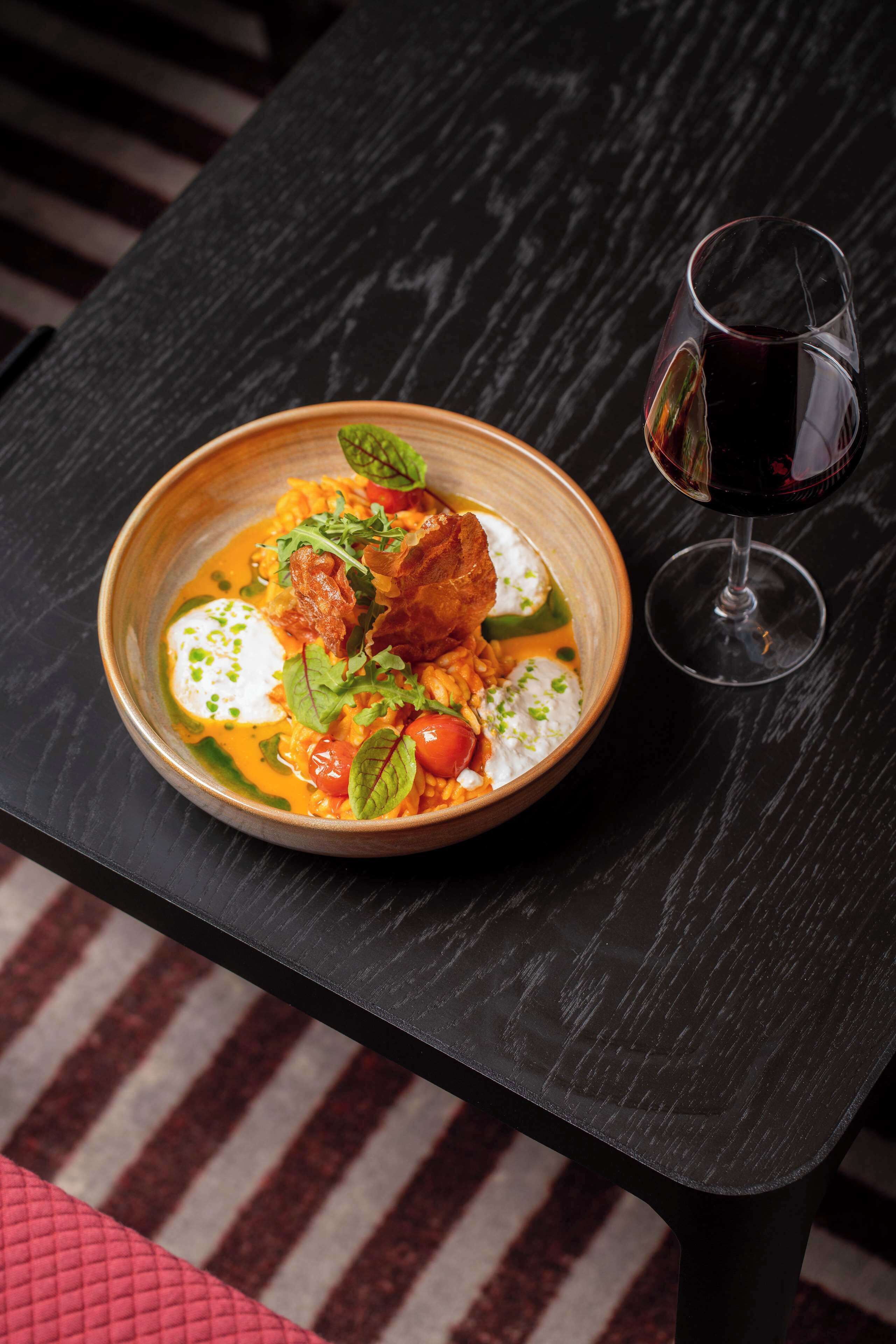 A bowl of food on a dark table with a glass of red wine beside it.