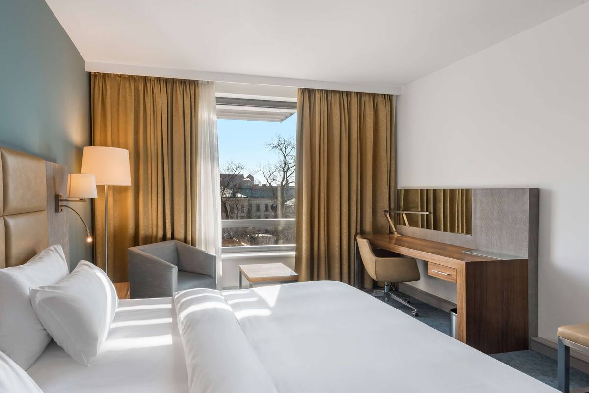 A hotel room with a bed, desk, chair, lamp, and large windows.