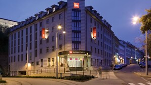 A modern hotel with a prominent 'ibis' sign, a central entrance with steps, and a parking area visible. - ibis Bratislava Centrum (Bratislava)