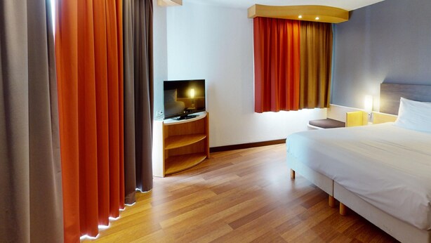 A hotel room with a bed, a TV on a stand, a nightstand, and curtains. - ibis Bratislava Centrum (Bratislava)