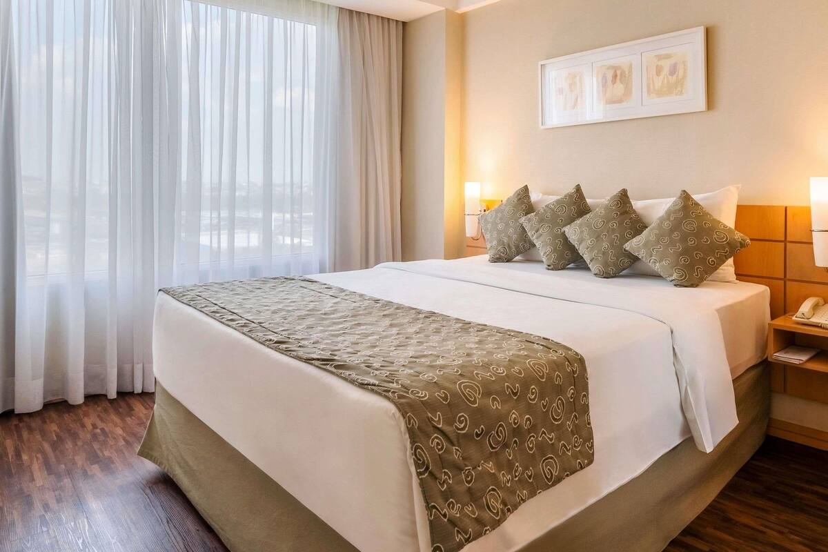 Una habitación de hotel con una cama grande, una mesita de noche con lámpara y una ventana con cortinas translúcidas.