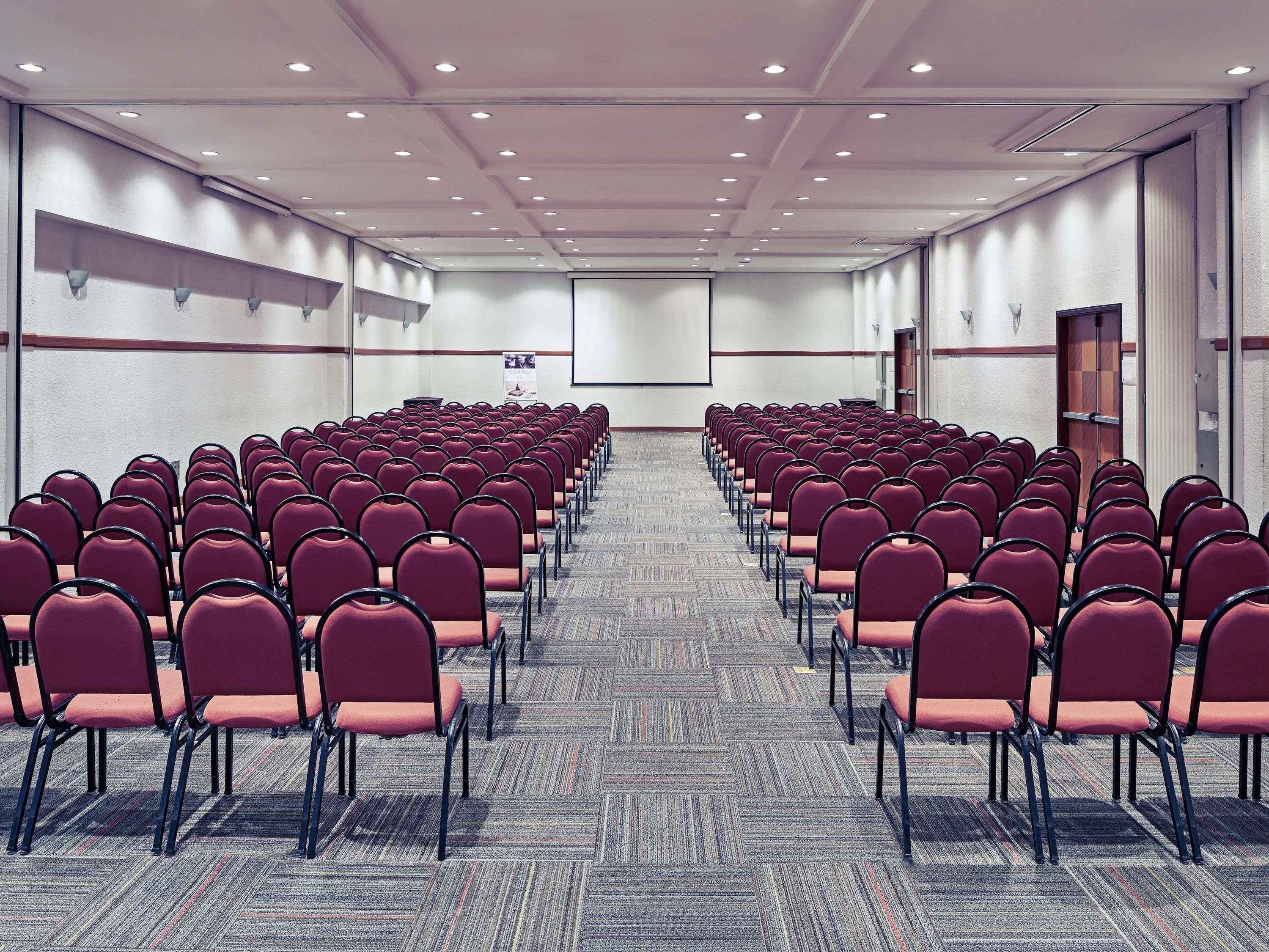 Un salón de conferencias con filas de sillas, piso alfombrado y una pantalla para proyector.