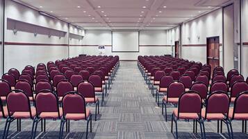Un salón de conferencias con filas de sillas, piso alfombrado y una pantalla para proyector.
