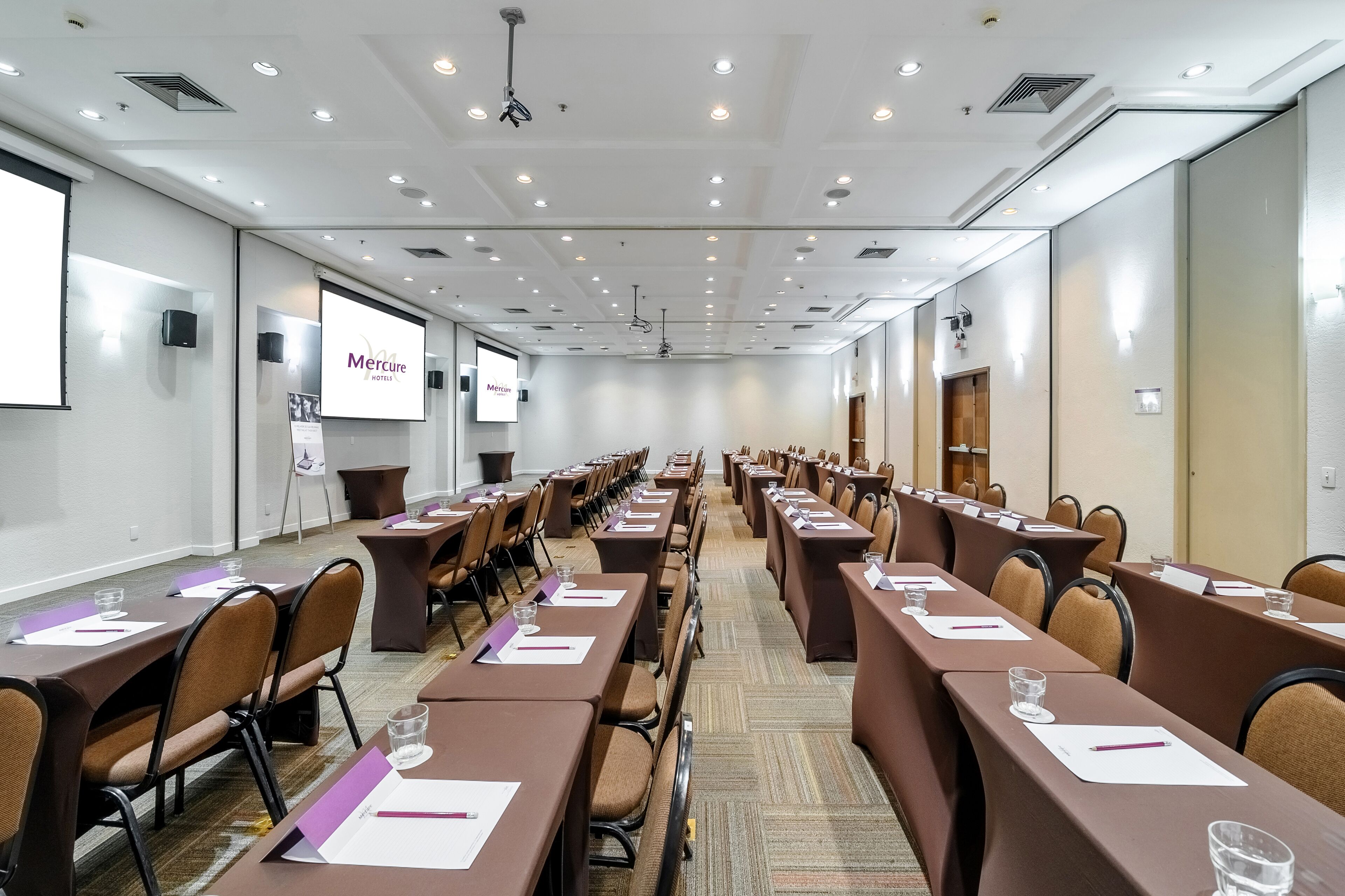 Un salón de conferencias con filas de mesas, sillas y una pantalla de proyección que muestra 'Mercure Hotels'.
