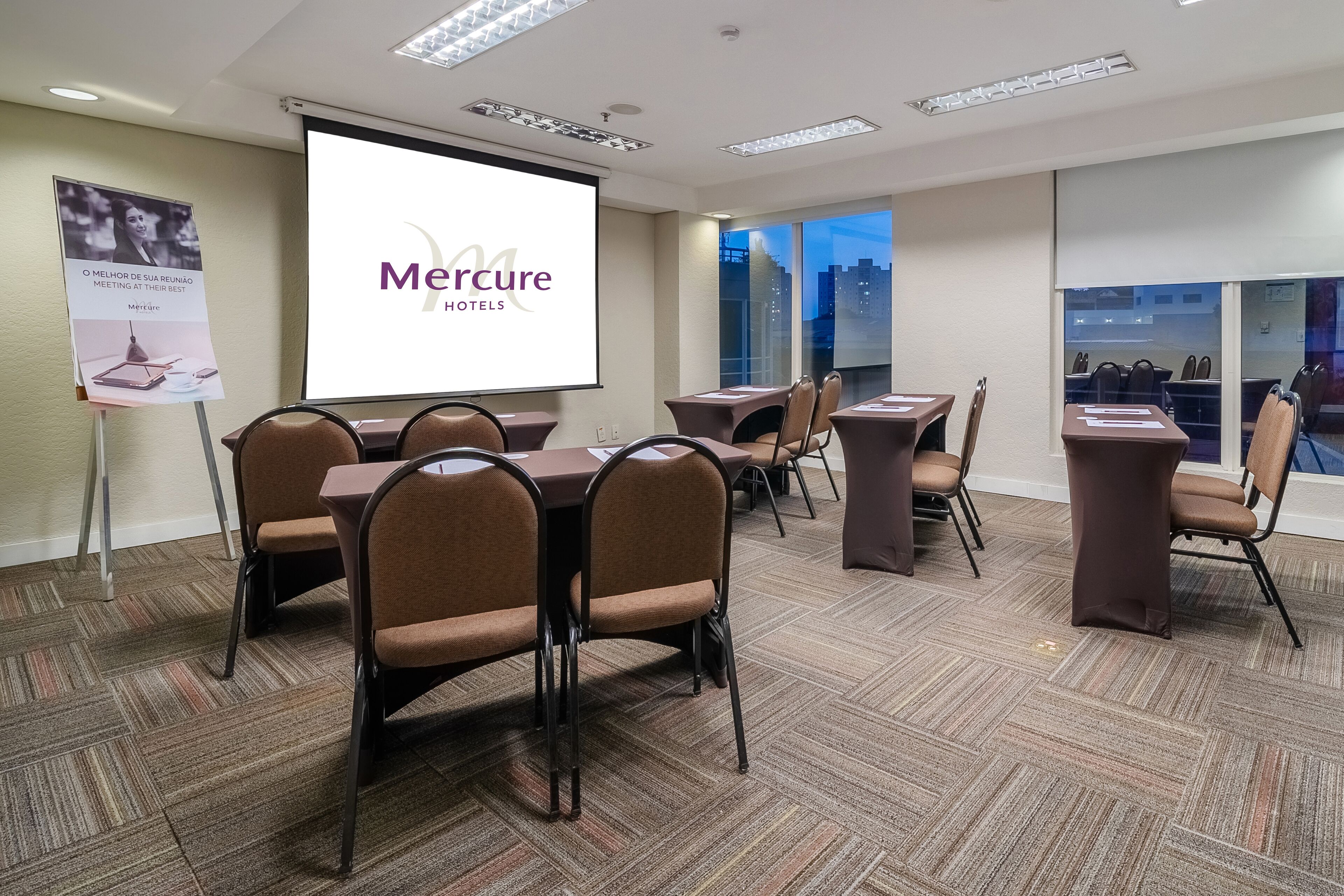 Une salle de conférence dotée d’un grand écran affichant le logo de la chaîne « Mercure Hotels », de plusieurs tables et chaises, d’un revêtement de sol en moquette et offrant une vue sur la ville grâce à de grandes fenêtres.