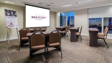 Une salle de conférence dotée d’un grand écran affichant le logo de la chaîne « Mercure Hotels », de plusieurs tables et chaises, d’un revêtement de sol en moquette et offrant une vue sur la ville grâce à de grandes fenêtres.