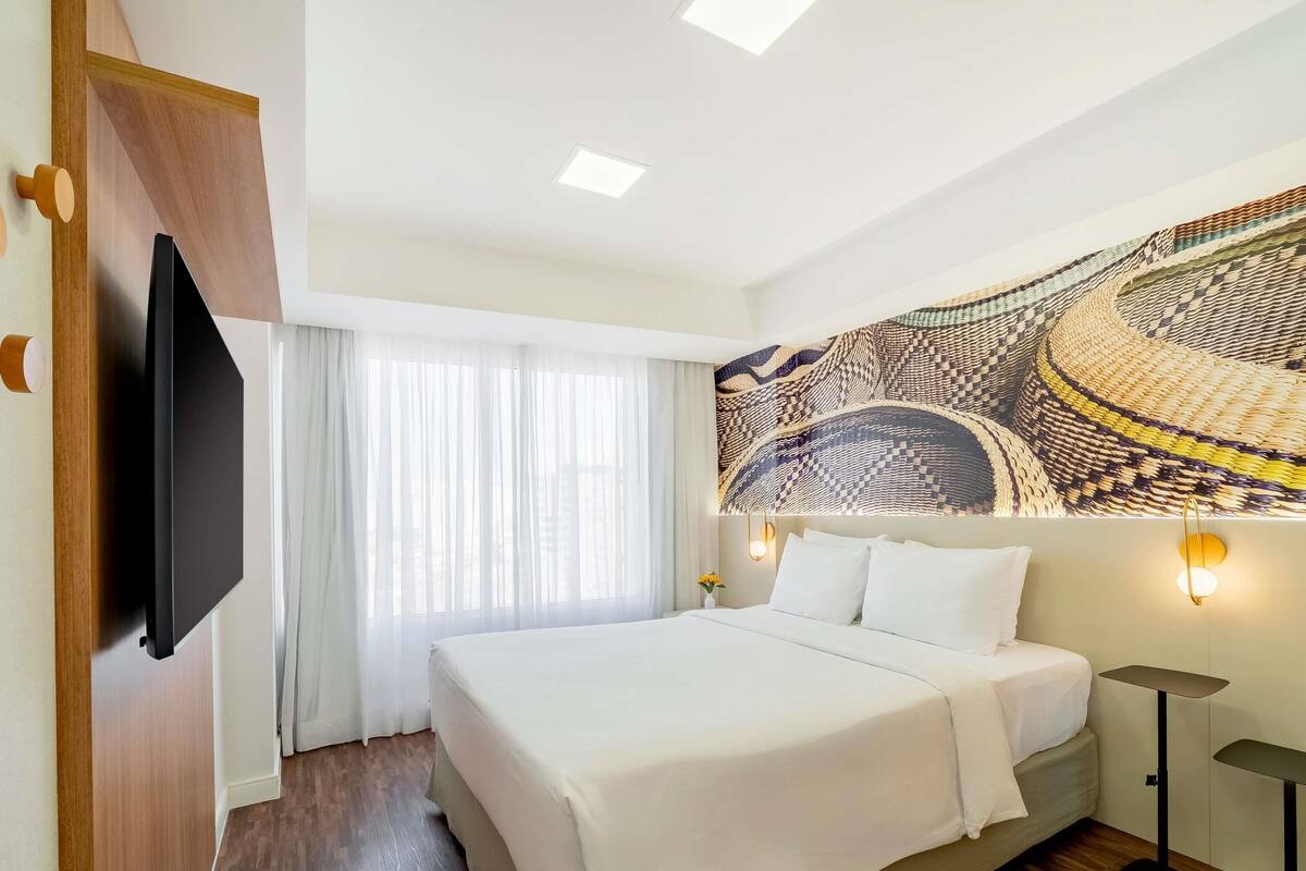 A hotel room with a bed, a flat-screen TV mounted on the wall, a large window with sheer curtains, a patterned wall mural, and a bedside table with a lamp.