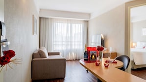Habitación de hotel con sofá, mesa de comedor con una botella de vino, florero con flores y cama.