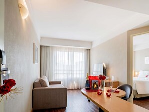 Habitación de hotel con sofá, mesa de comedor con una botella de vino, florero con flores y cama.