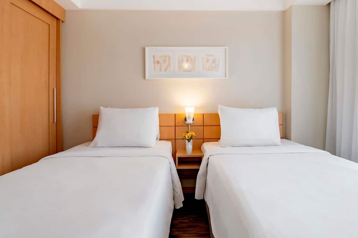 A hotel room with two beds, a wooden wardrobe, a nightstand with a lamp, and a framed picture on the wall.