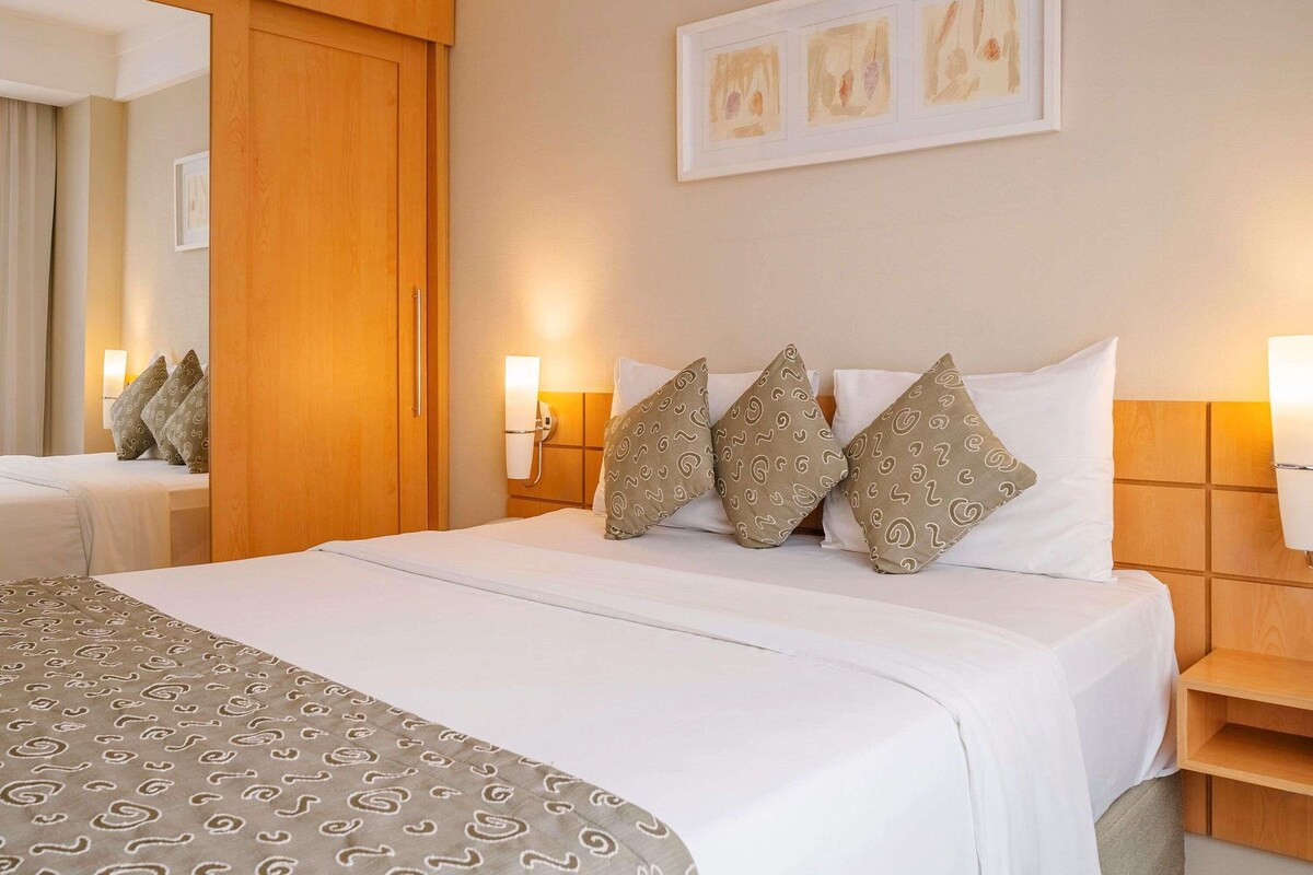 A hotel room with a large bed, a wooden wardrobe, a bedside table, and a wall-mounted light.