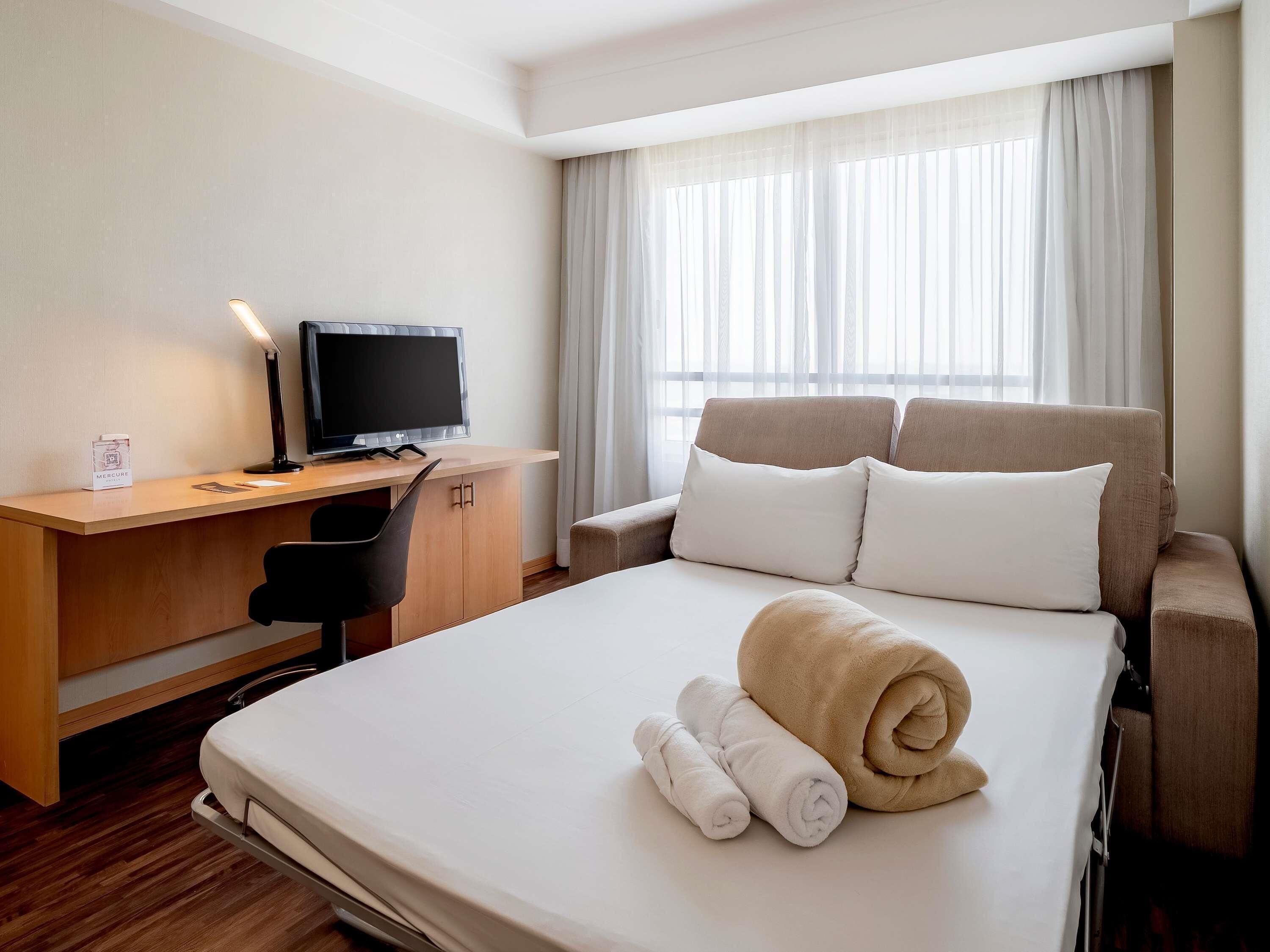 Ein Hotelzimmer mit Bett, Schreibtisch mit Computer, Stuhl und einem Fenster mit Vorhängen.