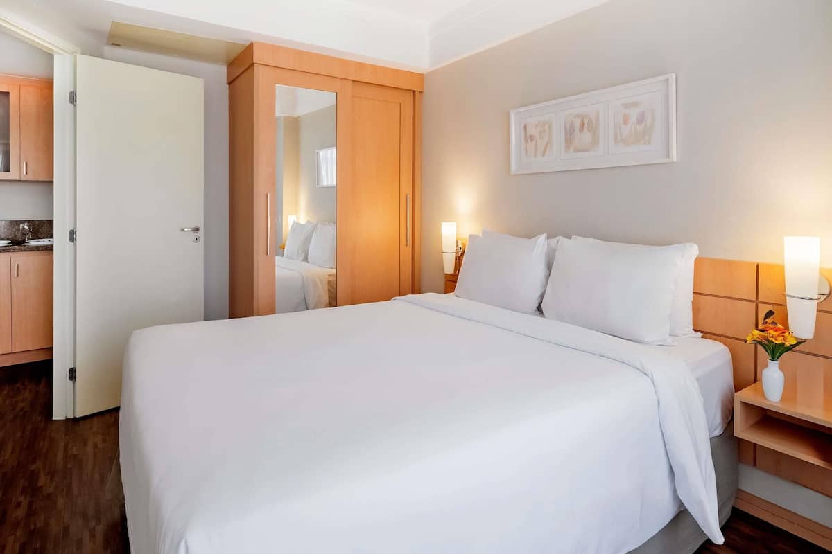 A hotel room with a large bed, wooden wardrobe, a small table with a vase, and a wall-mounted light fixture.