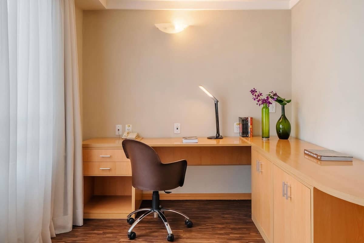 Ett hotellrum med ett skrivbord i trä, en stol, en bordslampa, en telefon, en vas med blommor och en bokhylla.