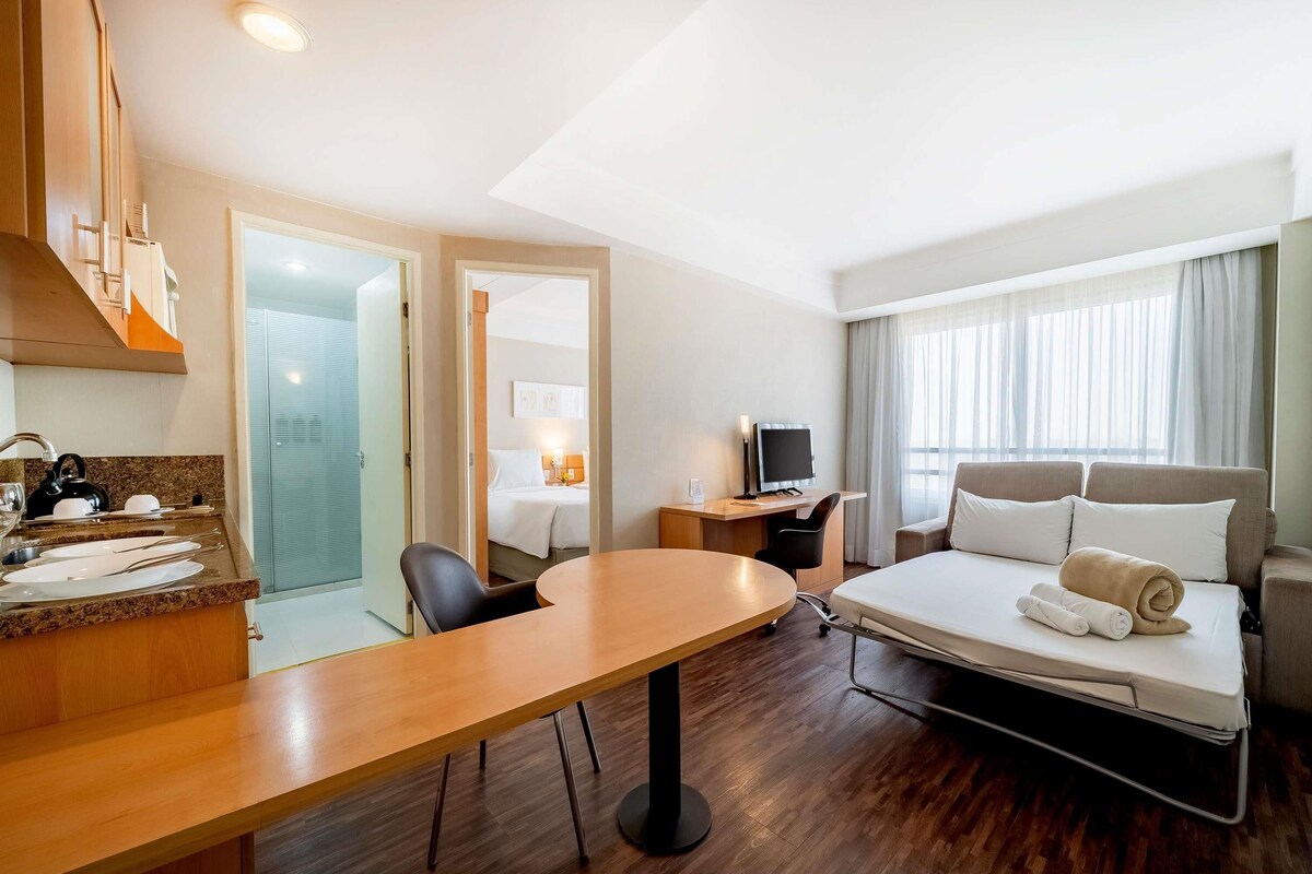 A hotel room with a bed, a desk with a computer, a chair, a table, and a kitchenette.