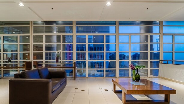 A spacious living area with a large window, a brown leather sofa, a wooden coffee table, and a vase with flowers. - Mercure Guarulhos Aeroporto Hotel (Guarulhos)