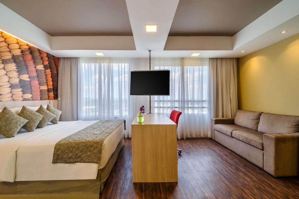 A hotel room with a bed, a desk with a TV, a sofa, and a stone wall.
