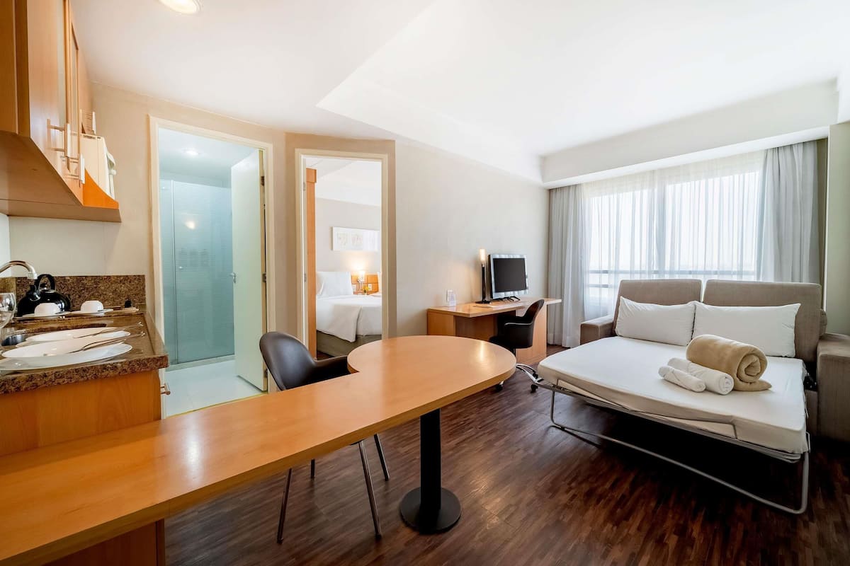 A hotel room with a bed, a desk, a chair, a TV, and a dining area with a table and chairs.