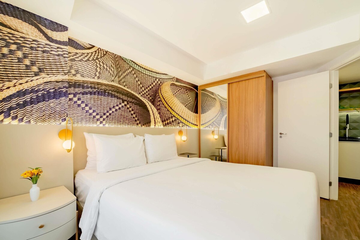 Habitación de hotel con una cama grande, una mesita de noche con un jarrón de flores, un tapiz en la pared y un armario de madera.