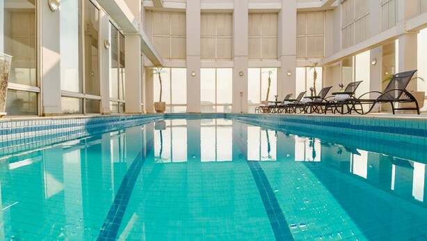 An indoor swimming pool with clear blue water, sunlit interior, and lounge chairs along the sides. - Mercure Guarulhos Aeroporto Hotel (Guarulhos)