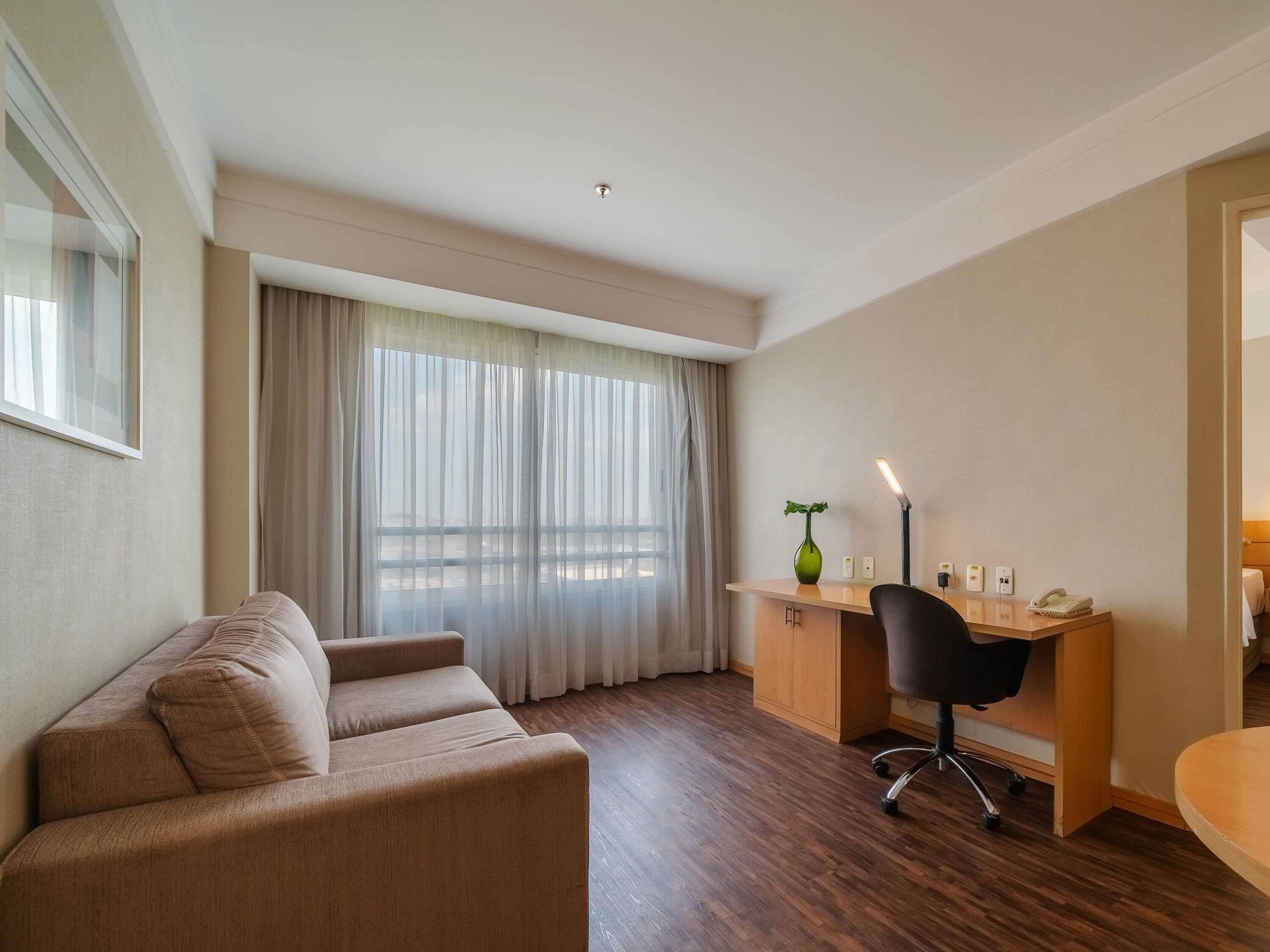 A modern hotel room with a brown leather sofa, a wooden desk with a chair, a green vase, and a large window with sheer curtains.