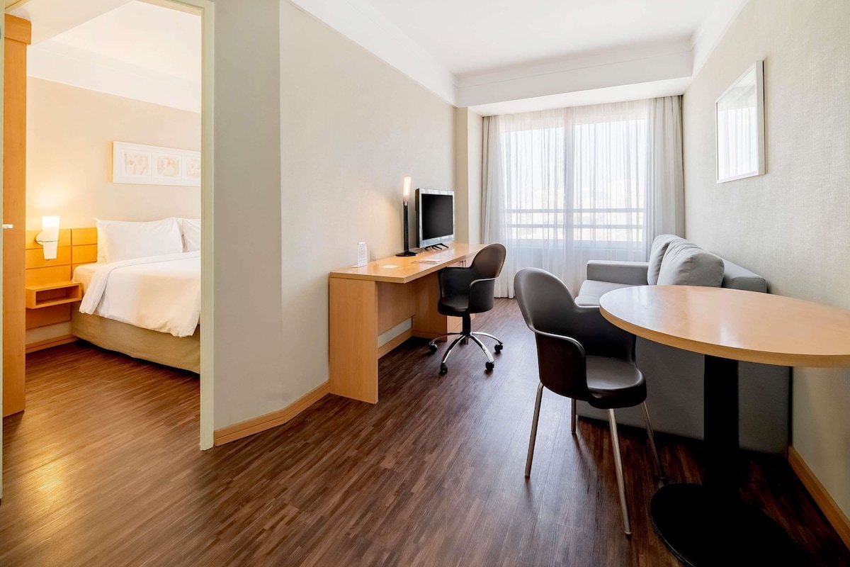 Habitación de hotel con cama, escritorio con computadora, silla, sofá y ventana con cortinas.