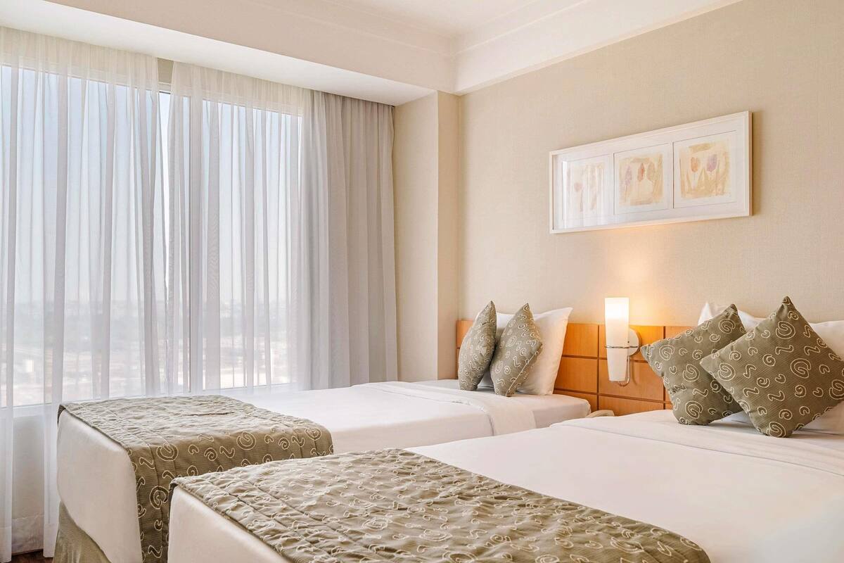 A hotel room with two beds, a large window with sheer curtains, a wall-mounted light, and a framed picture above the beds.