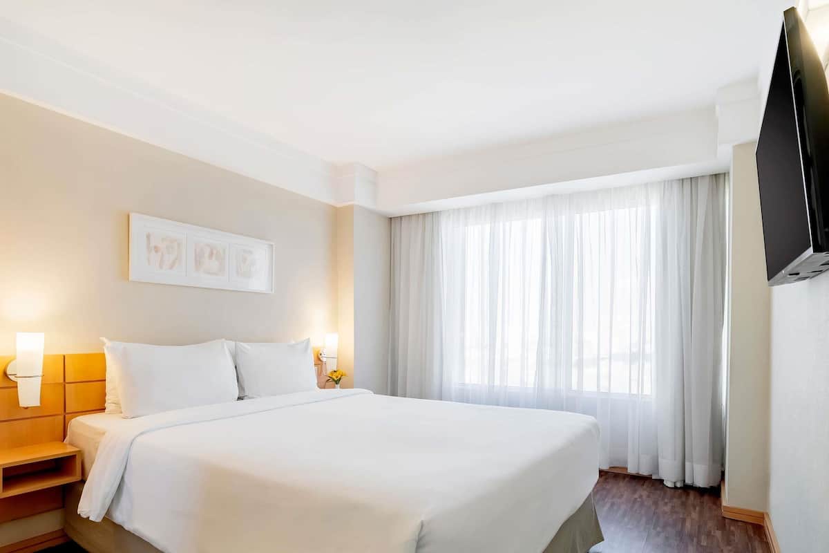 A hotel room with a large bed, bedside lamps, a flat-screen TV, and a window with sheer curtains.